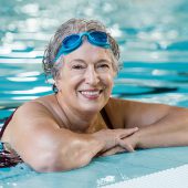 Mature woman wearing swim goggles at swimming pool. Fit active senior woman enjoying retirement standing in swimming pool and looking at camera. Happy senior healthy old woman enjoying active lifestyle.